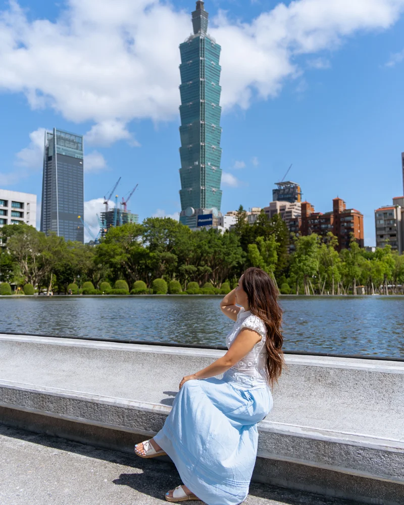 Woman sitting in front of Reflecting Pool at National Dr. Sun Yat-sen Memorial Hall with Taipei 101 in the background.